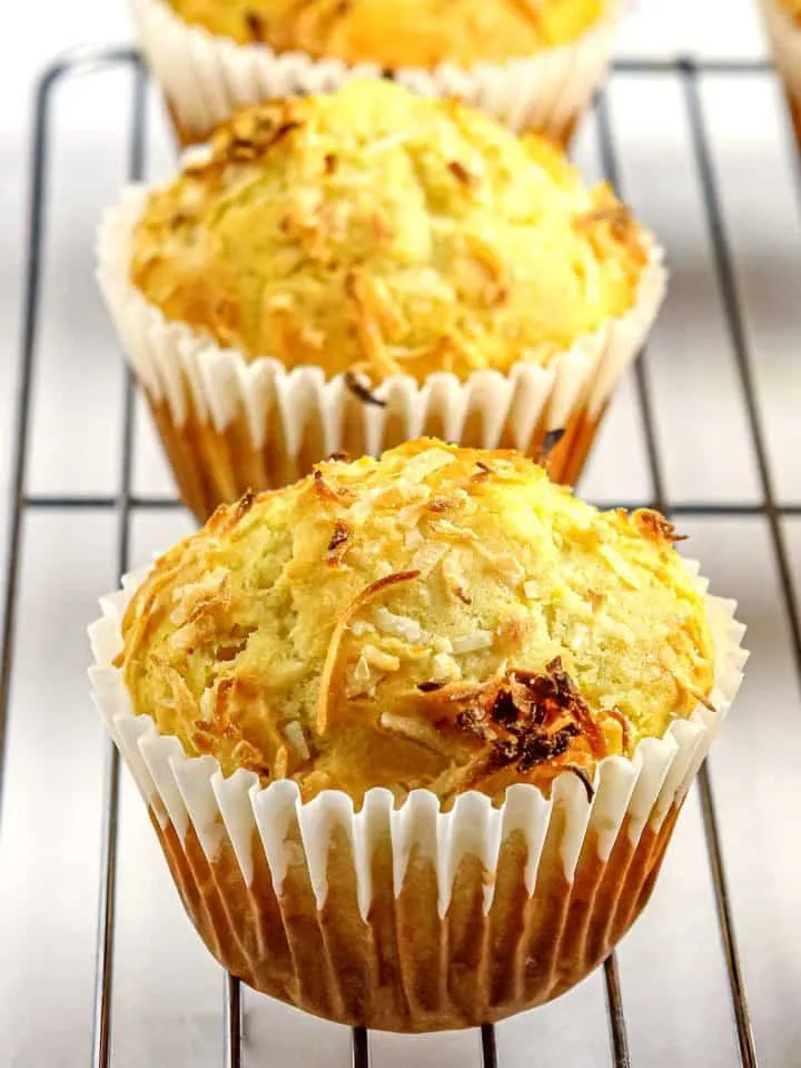 Three coconut-topped muffins in white liners cooling on a metal rack against a white background.