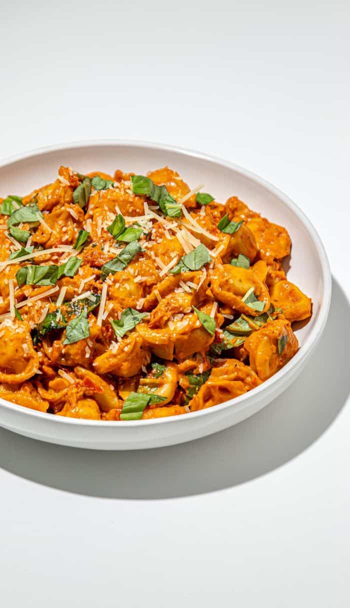 White bowl of tortellini pasta in creamy tomato sauce, topped with shredded cheese and fresh basil leaves on white background.