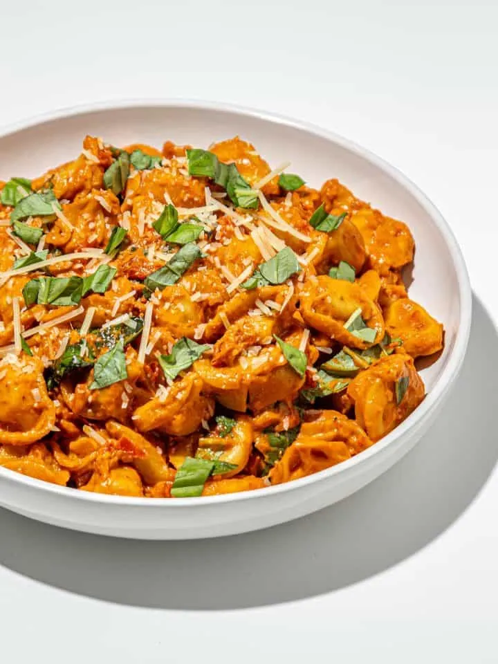 White bowl of tortellini pasta in creamy tomato sauce, topped with shredded cheese and fresh basil leaves on white background.
