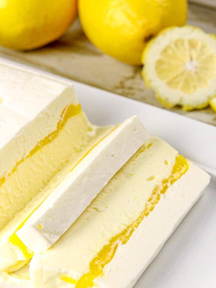 Layered lemon mousse dessert with a yellow lemon curd center, two fresh whole lemons, and one lemon slice on a plate.