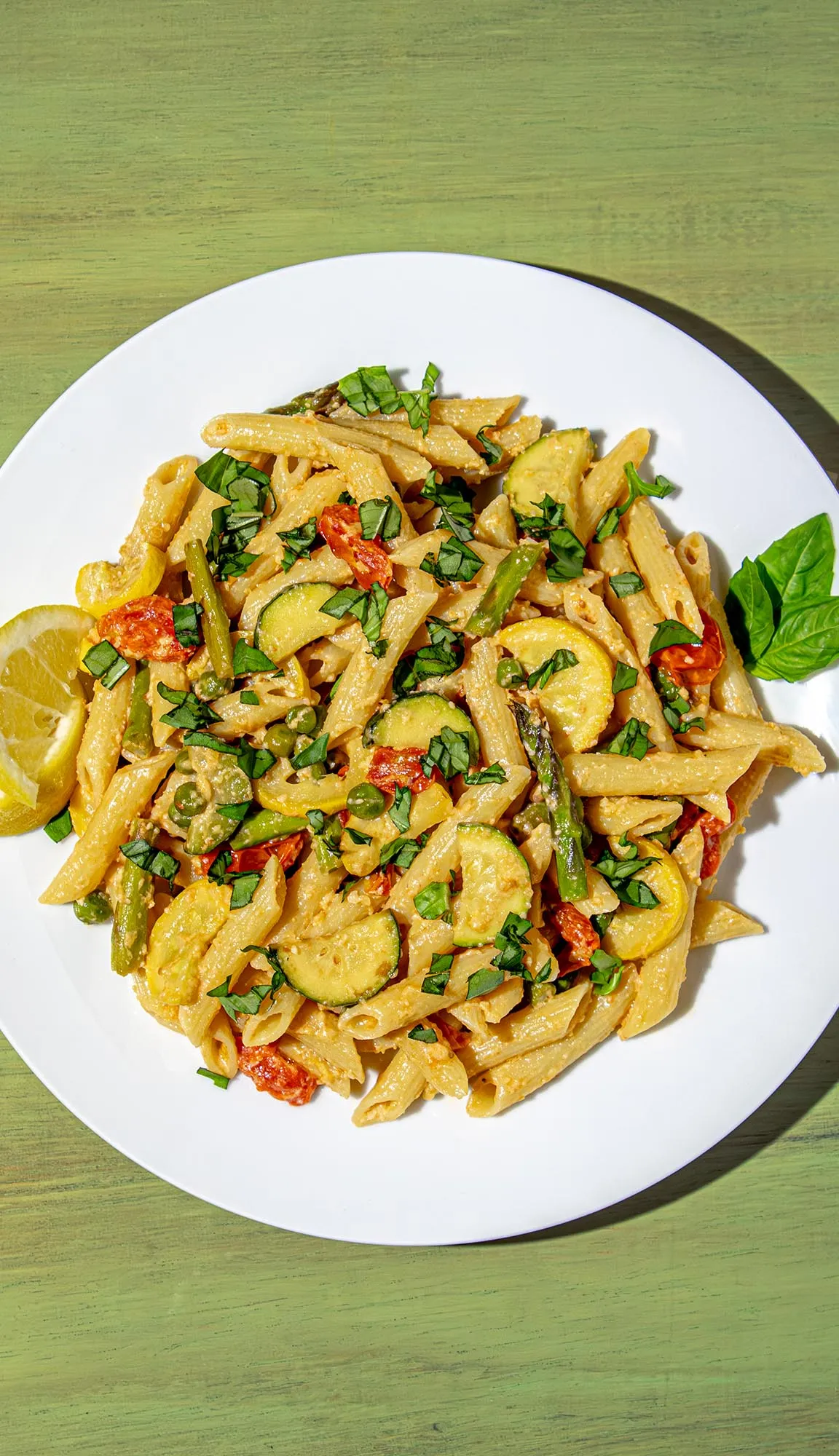 Penne pasta with zucchini, yellow squash, asparagus, cherry tomatoes, peas, and chopped basil on a white plate with lemon wedges and a basil leaf.