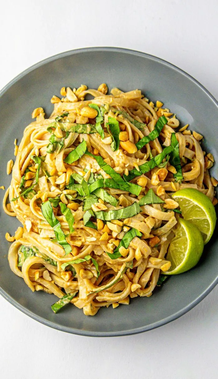 Noodles in creamy peanut sauce garnished with chopped peanuts, fresh basil, and two lime wedges in a gray bowl on a white surface.