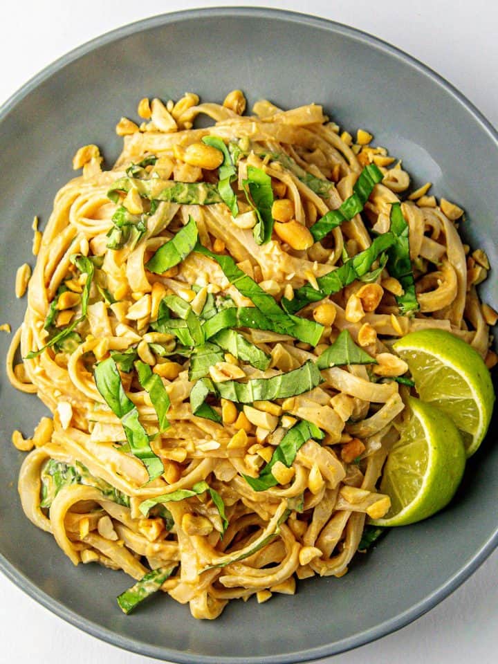 Noodles in creamy peanut sauce garnished with chopped peanuts, fresh basil, and two lime wedges in a gray bowl on a white surface.