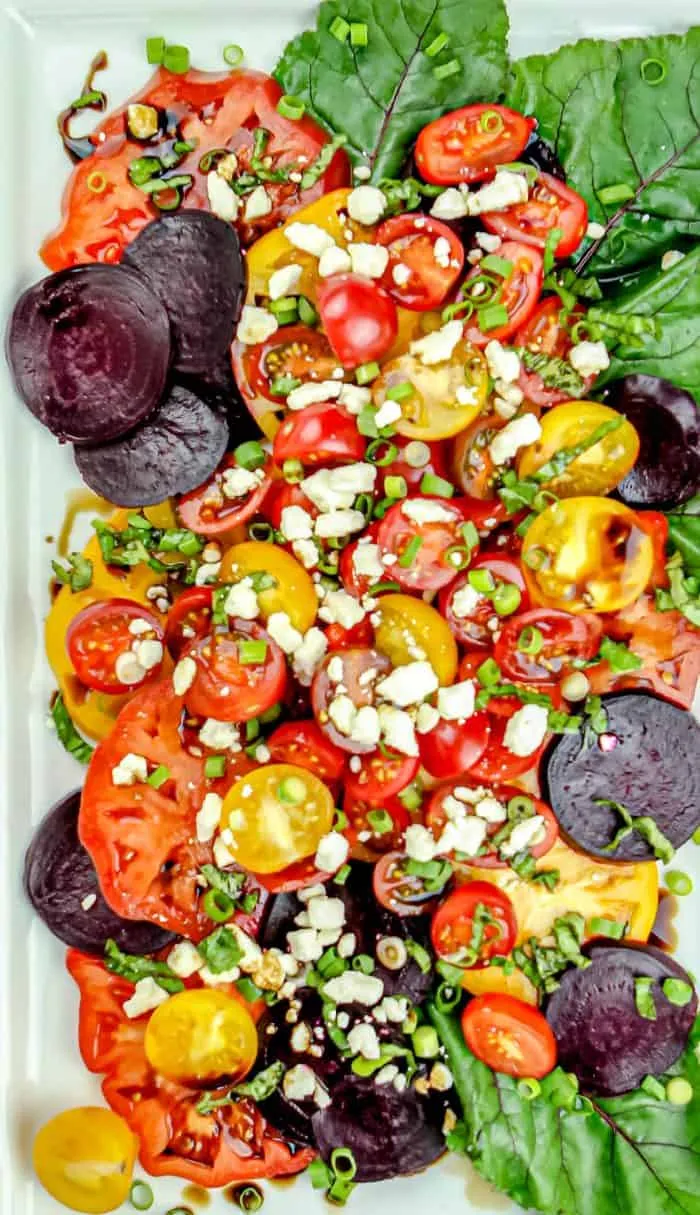 Colorful salad with sliced red and yellow tomatoes, beet slices, crumbled cheese, chopped green onions, and leafy greens on a white plate.
