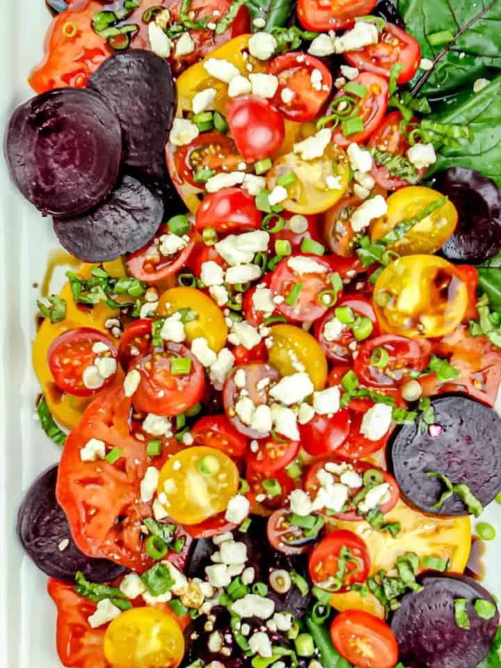 Colorful salad with sliced red and yellow tomatoes, beet slices, crumbled cheese, chopped green onions, and leafy greens on a white plate.