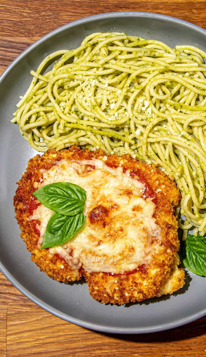 Breaded chicken parmesan with melted cheese and basil leaf, served with spaghetti coated in green pesto sauce on a gray plate.