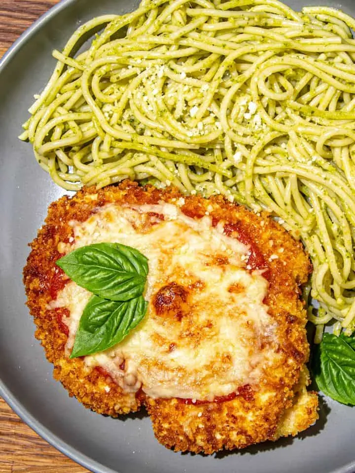 Breaded chicken parmesan with melted cheese and basil leaf, served with spaghetti coated in green pesto sauce on a gray plate.