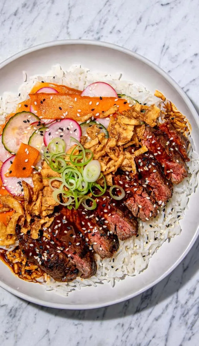 Grilled steak slices with red thai curry sauce, sesame seeds, fried onions, pickled cucumber and radish, carrot ribbons on white rice in a white bowl.