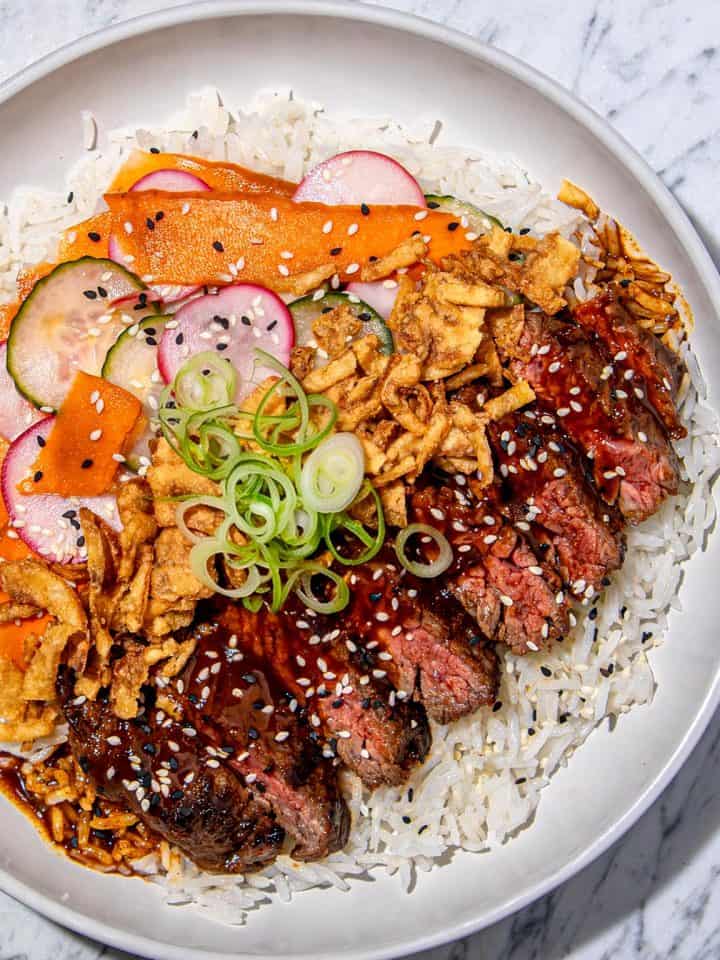 Grilled steak slices with red thai curry sauce, sesame seeds, fried onions, pickled cucumber and radish, carrot ribbons on white rice in a white bowl.