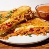 Grilled sandwich with melted cheese, shredded chicken, and herbs on a white plate, with a small bowl of butter chicken sauce on the side.