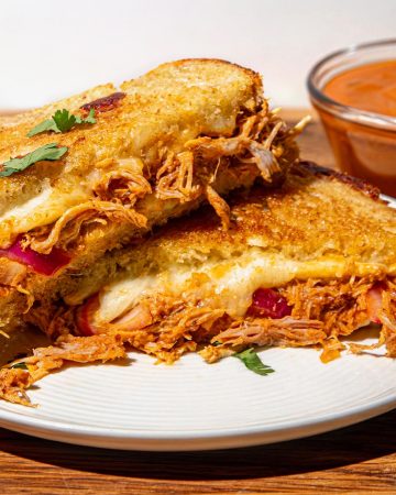 Grilled sandwich with melted cheese, shredded chicken, and herbs on a white plate, with a small bowl of butter chicken sauce on the side.