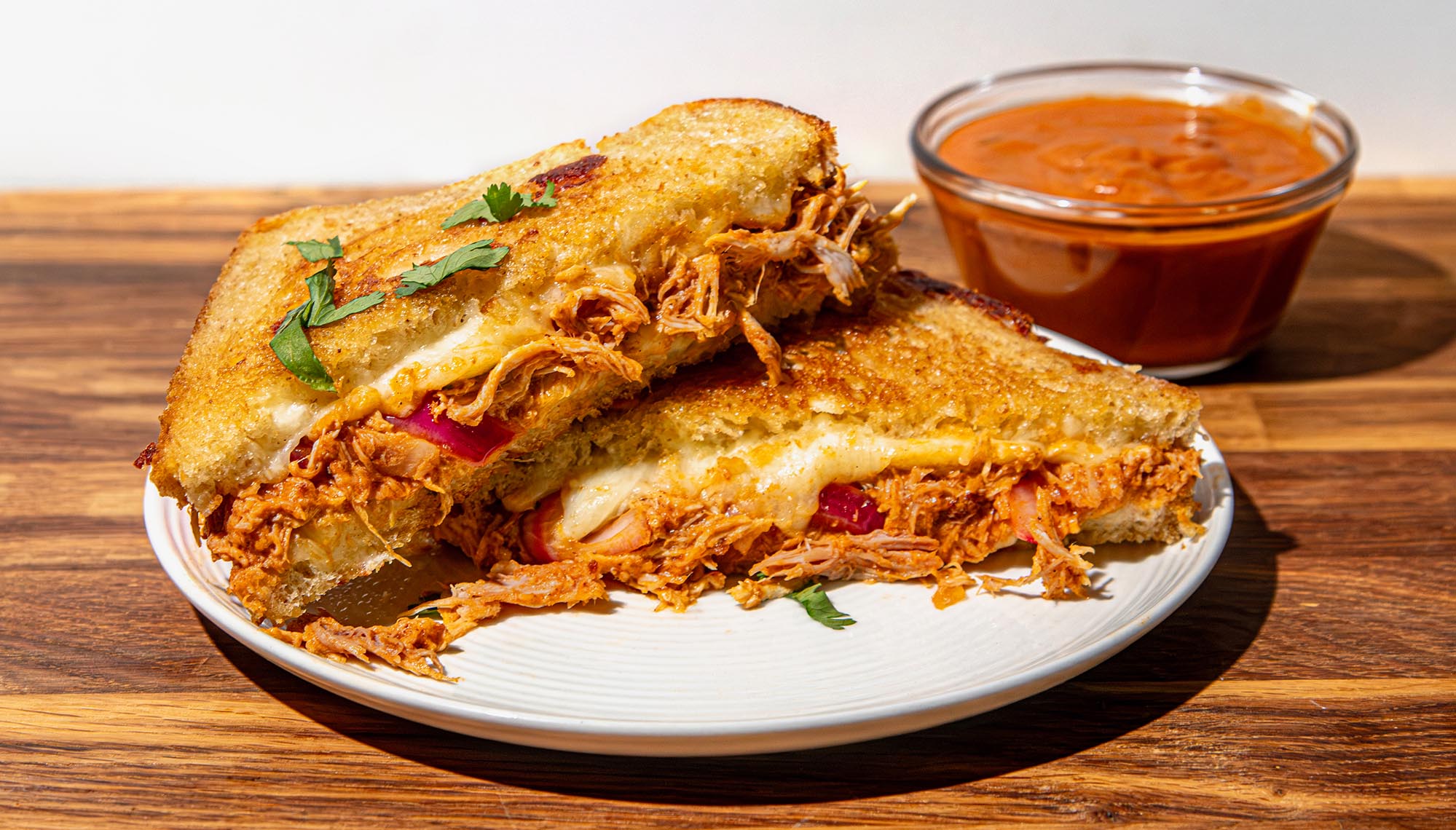 Grilled sandwich with melted cheese, shredded chicken, and herbs on a white plate, with a small bowl of butter chicken sauce on the side.
