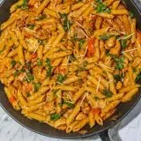 Pasta with creamy sauce, chicken pieces, diced red bell peppers, sprinkled cheese, and chopped herbs in a black skillet on a marble surface.