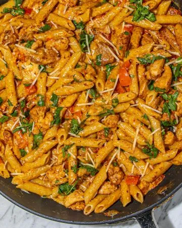 Pasta with creamy sauce, chicken pieces, diced red bell peppers, sprinkled cheese, and chopped herbs in a black skillet on a marble surface.