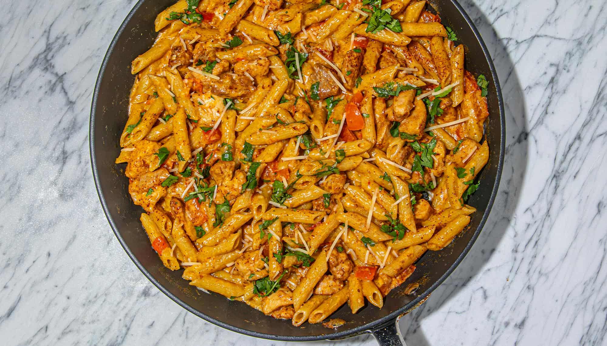 Pasta with creamy sauce, chicken pieces, diced red bell peppers, sprinkled cheese, and chopped herbs in a black skillet on a marble surface.