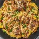Pasta with seared chicken strips, shredded cheese, bacon bits, and chopped green onions in a black pan on a marble surface.