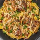 Pasta with seared chicken strips, shredded cheese, bacon bits, and chopped green onions in a black pan on a marble surface.
