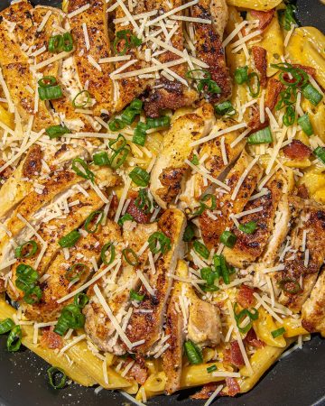 Pasta with seared chicken strips, shredded cheese, bacon bits, and chopped green onions in a black pan on a marble surface.