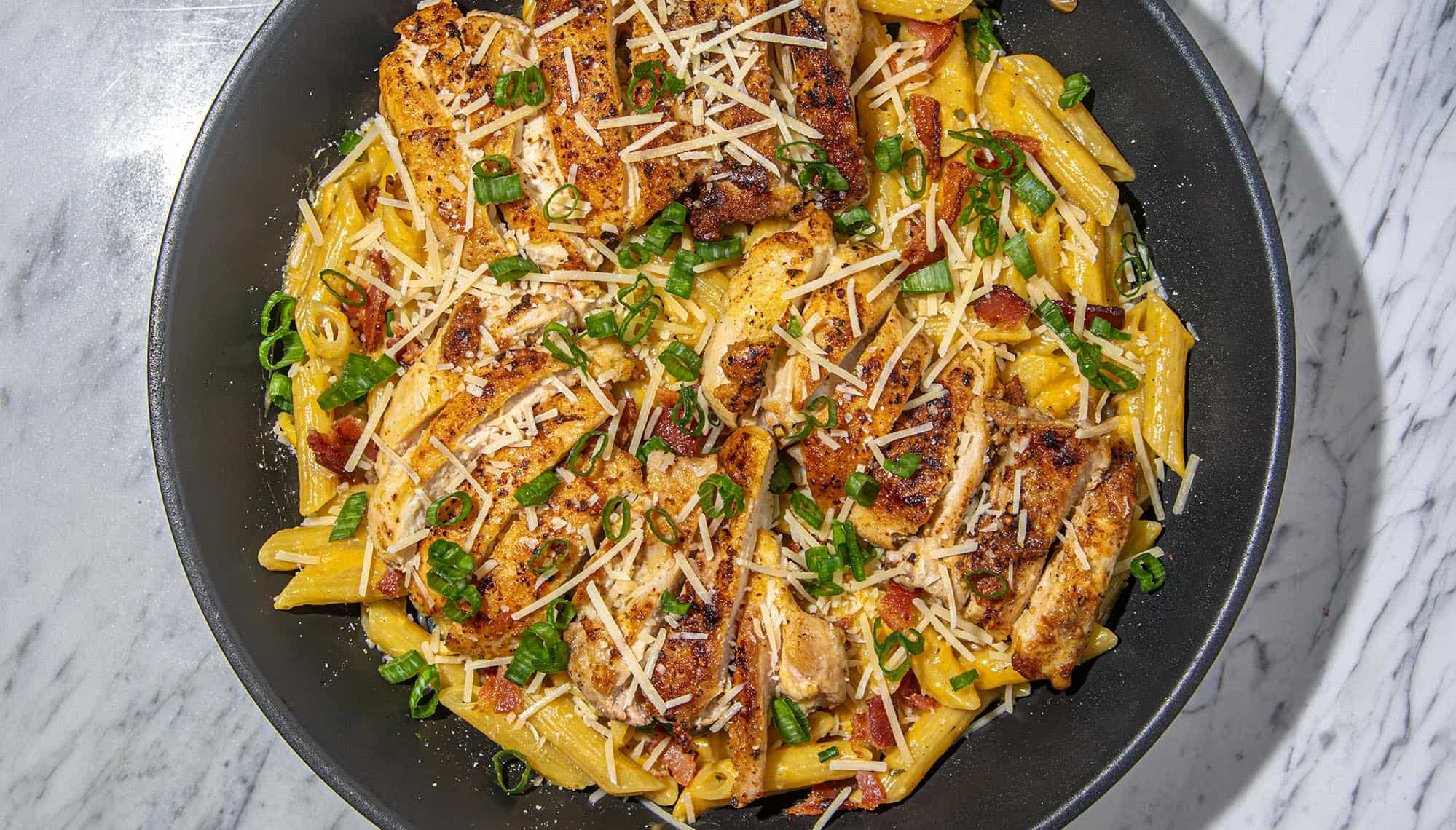 Pasta with seared chicken strips, shredded cheese, bacon bits, and chopped green onions in a black pan on a marble surface.