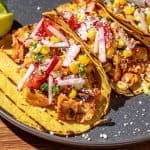 Grilled chicken tacos with corn salsa, radish, tomato, and crumbled cheese on yellow corn tortillas with lime wedges on gray plate.