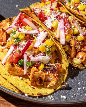 Grilled chicken tacos with corn salsa, radish, tomato, and crumbled cheese on yellow corn tortillas with lime wedges on gray plate.
