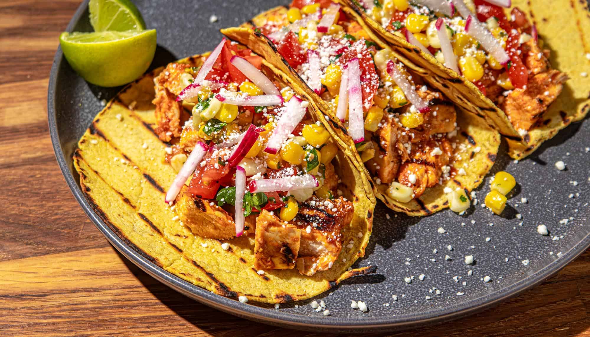 Grilled chicken tacos with corn salsa, radish, tomato, and crumbled cheese on yellow corn tortillas with lime wedges on gray plate.