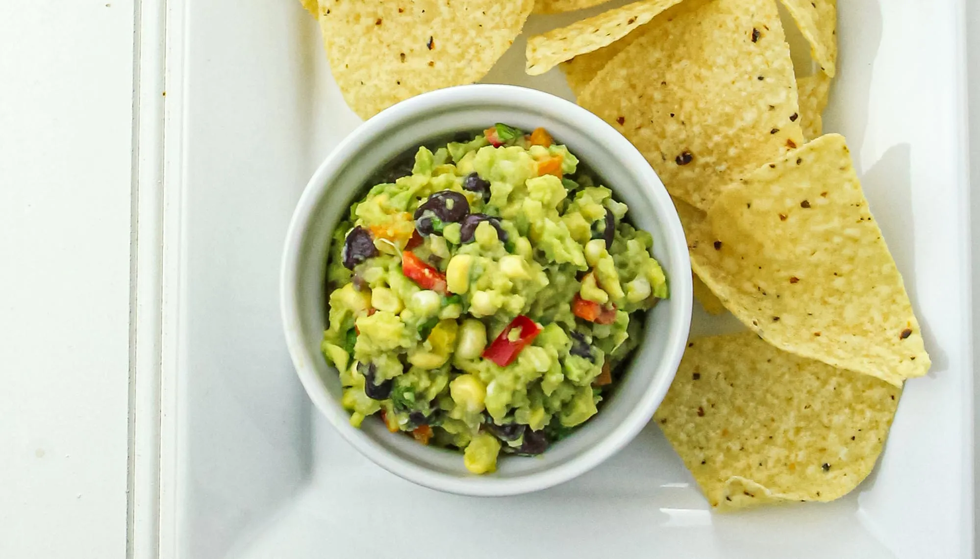 Bowl of guacamole with black beans, corn, and red peppers served with tortilla chips on a white plate.