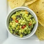 Bowl of guacamole with black beans, corn, and red pepper next to yellow corn tortilla chips on a white plate.