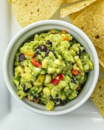 Bowl of guacamole with black beans, corn, and red pepper next to yellow corn tortilla chips on a white plate.