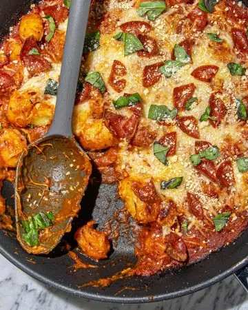 Gnocchi baked with tomato sauce, melted cheese, pepperoni slices, grated parmesan, and fresh basil on a marble surface.