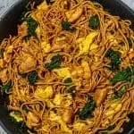 Stir-fried noodles with chicken, scrambled eggs, and kale in a black skillet on a marble surface.