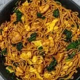 Stir-fried noodles with chicken, scrambled eggs, and kale in a black skillet on a marble surface.