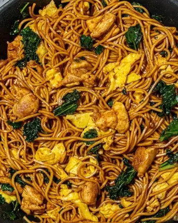 Stir-fried noodles with chicken, scrambled eggs, and kale in a black skillet on a marble surface.