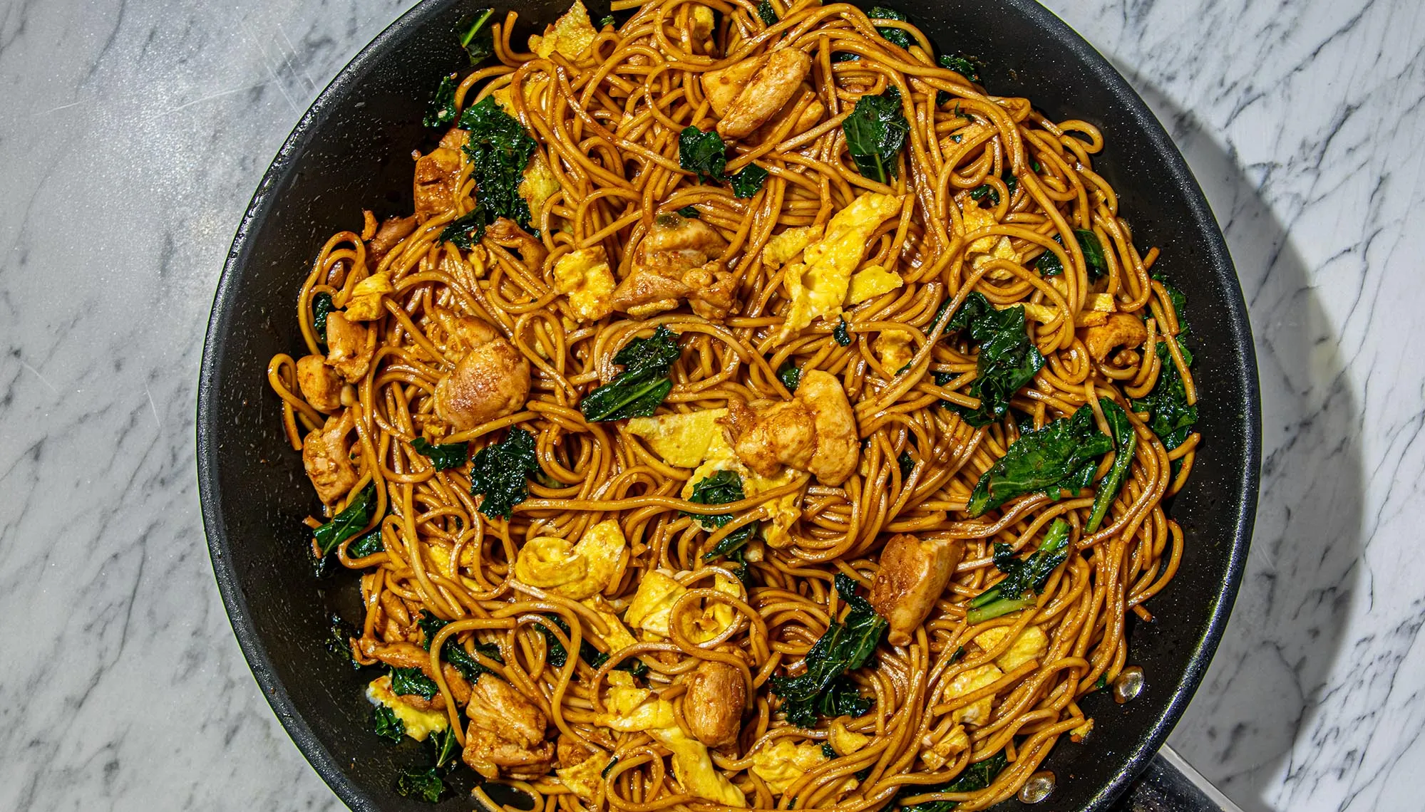 Stir-fried noodles with chicken, scrambled eggs, and kale in a black skillet on a marble surface.