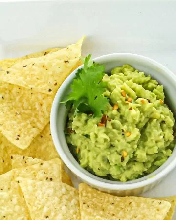 Guacamole garnished with cilantro and red pepper flakes in a white bowl, surrounded by yellow corn tortilla chips on a white plate.
