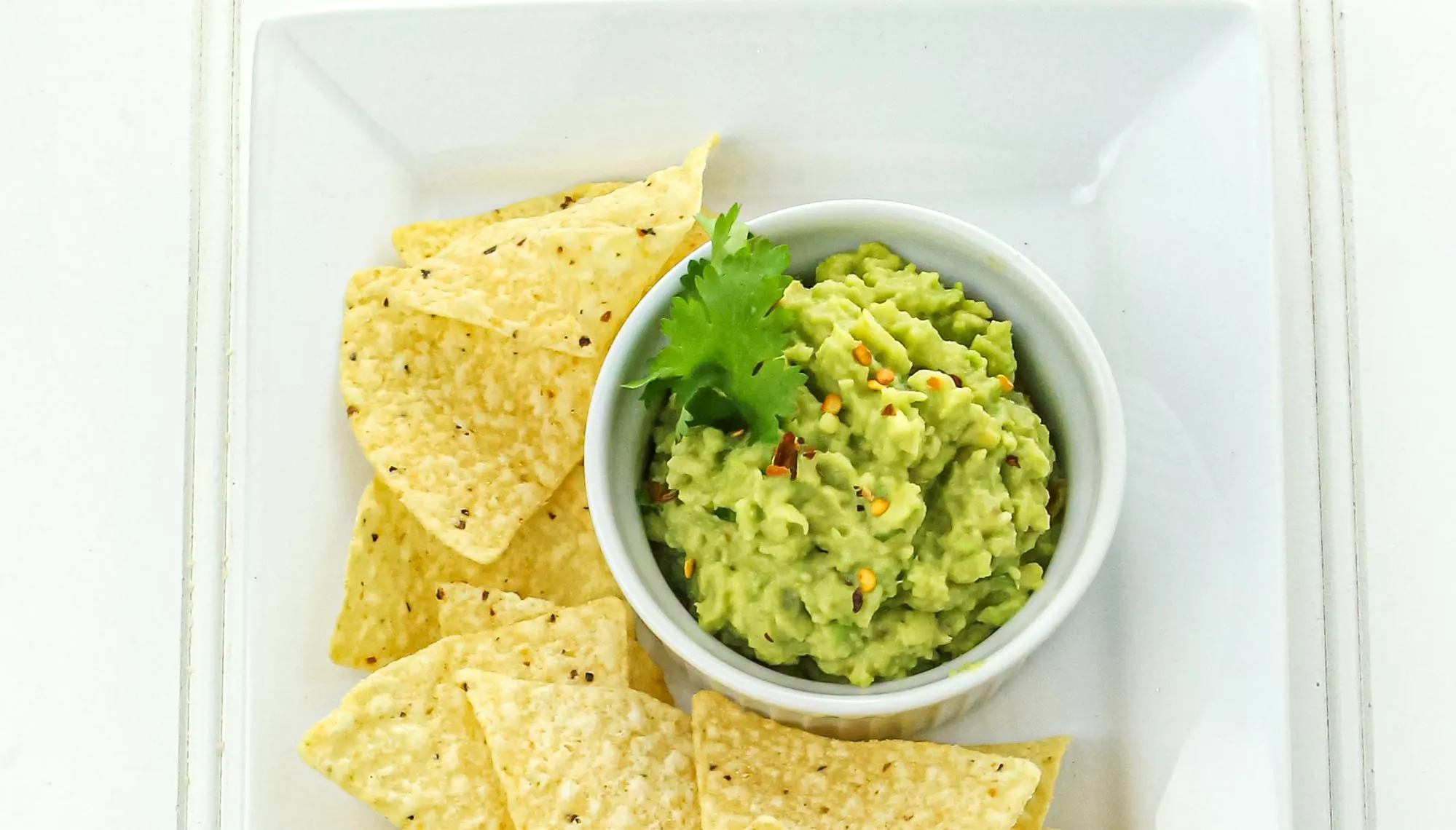 Guacamole garnished with cilantro and red pepper flakes in a white bowl, surrounded by yellow corn tortilla chips on a white plate.