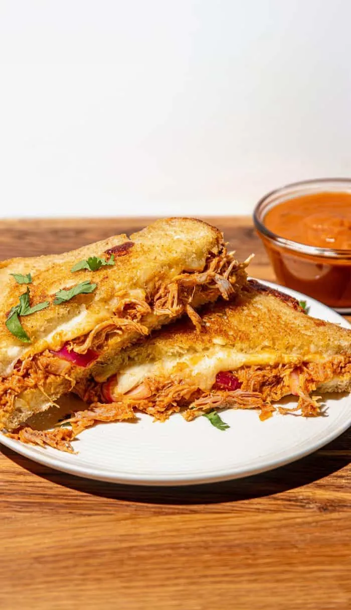 Grilled cheese sandwich with shredded butter chicken and cilantro, served on a white plate with a bowl of butter chicken sauce.