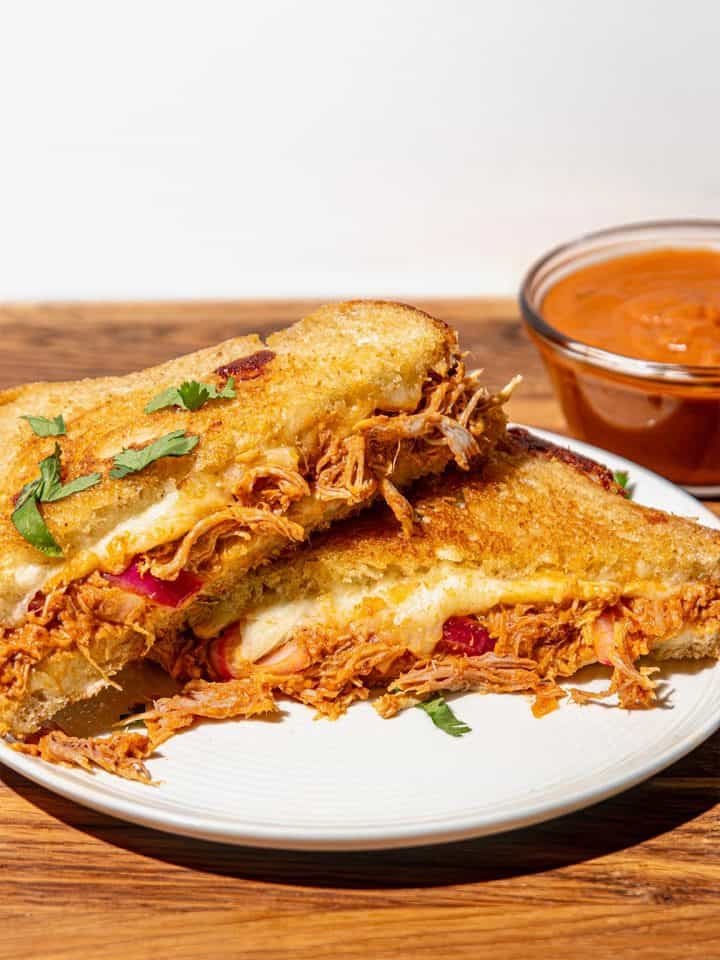 Grilled cheese sandwich with shredded butter chicken and cilantro, served on a white plate with a bowl of butter chicken sauce.