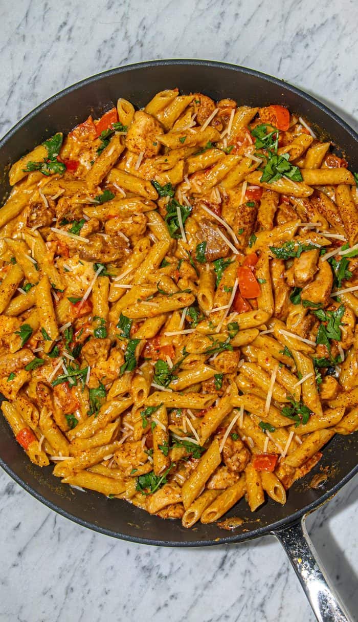 Penne pasta with grilled chicken, diced red peppers, fresh cilantro, and grated cheese in a creamy sauce in a black skillet on marble surface.
