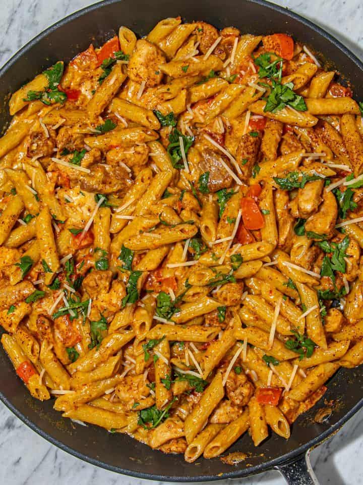 Penne pasta with grilled chicken, diced red peppers, fresh cilantro, and grated cheese in a creamy sauce in a black skillet on marble surface.