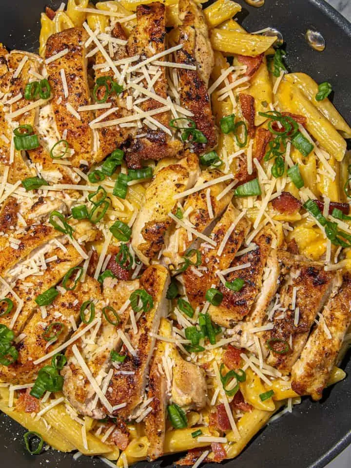 Seared chicken strips over creamy penne pasta with bacon, sprinkled with chopped green onions and shredded Parmesan cheese in a black skillet.
