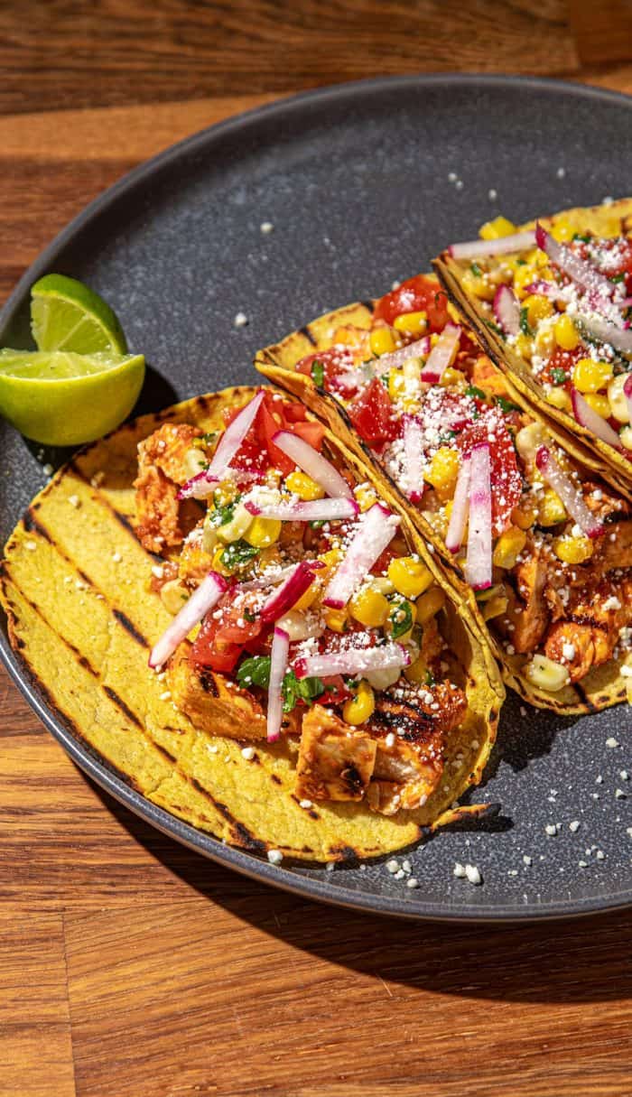 Grilled chicken tacos with corn, radish, tomato, and cheese on a dark plate with lime wedges on wooden table.