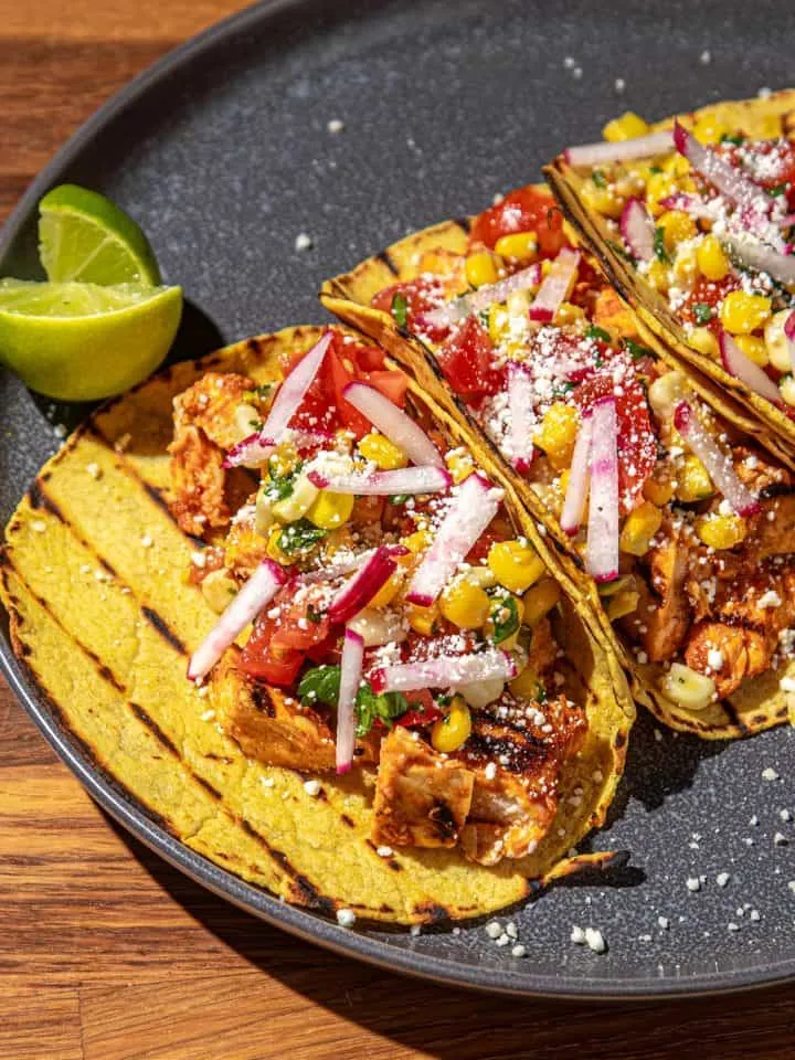 Grilled chicken tacos with corn, radish, tomato, and cheese on a dark plate with lime wedges on wooden table.