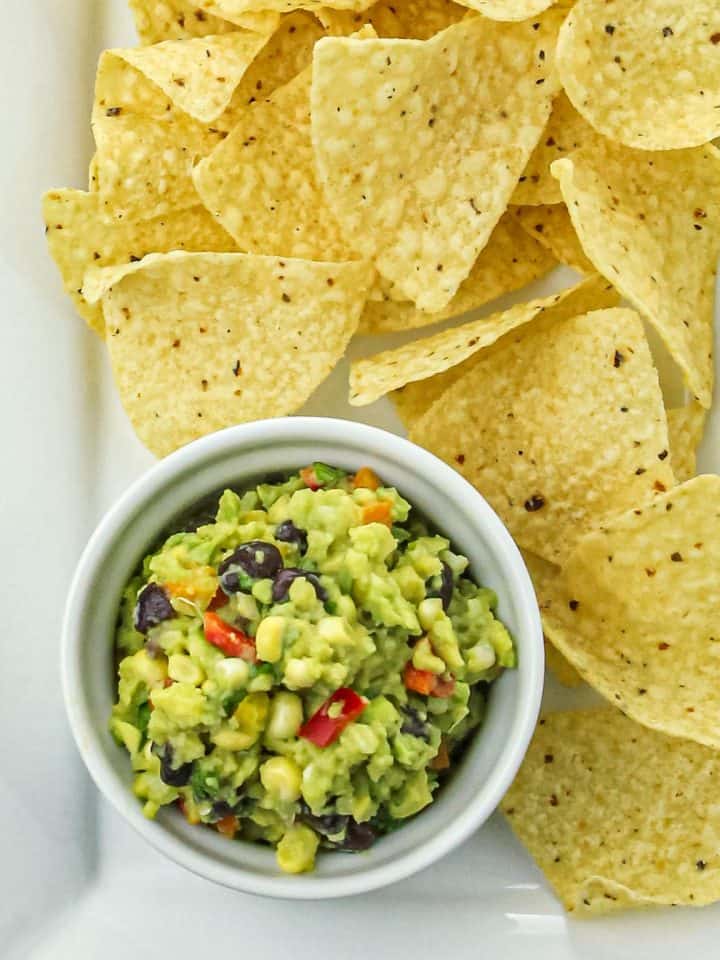 Corn tortilla chips served with a bowl of guacamole mixed with black beans, corn, and diced red peppers on a white plate.