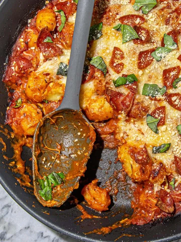Skillet with pepperoni pizza topped baked gnocchi with basil and parmesan, next to a large serving spoon.