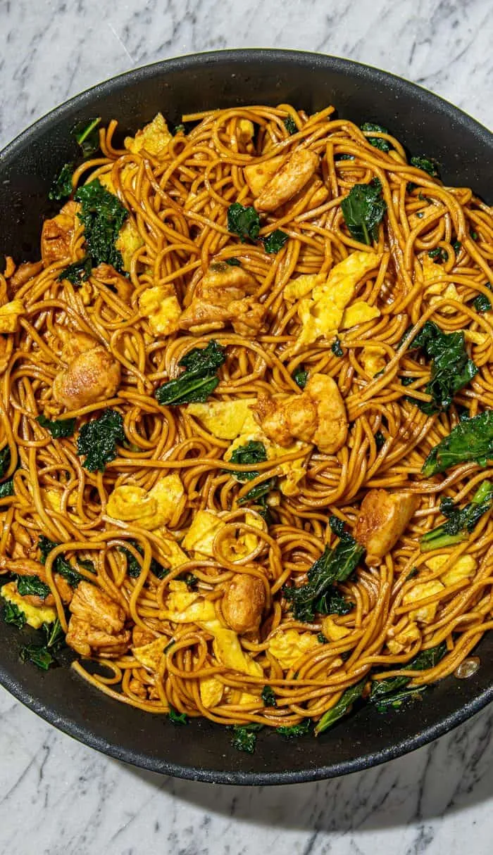 Stir-fry with noodles, chicken pieces, scrambled eggs, and kale in a black pan on a marble surface.