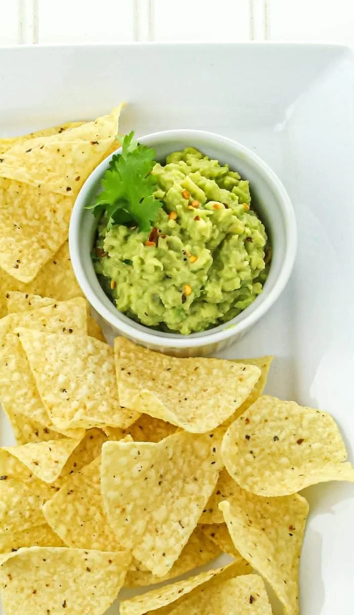 Yellow corn tortilla chips arranged around a bowl of guacamole garnished with cilantro and red pepper flakes on a white plate.
