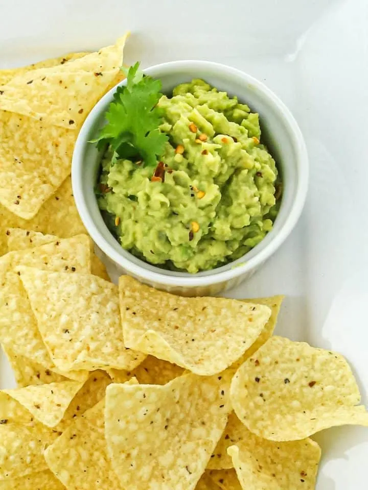 Yellow corn tortilla chips arranged around a bowl of guacamole garnished with cilantro and red pepper flakes on a white plate.