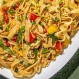 Stir-fried flat rice noodles with scrambled eggs, red and yellow bell peppers, green onions, and leafy greens on a white platter with a green textured cloth.