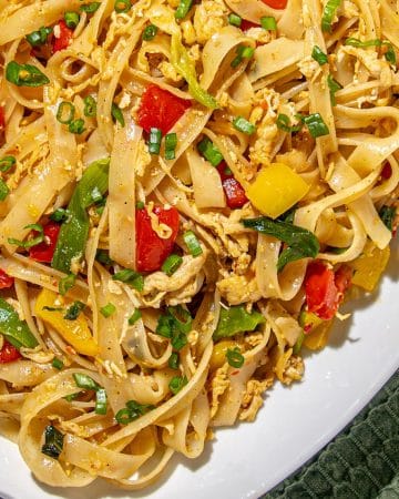 Stir-fried flat rice noodles with scrambled eggs, red and yellow bell peppers, green onions, and leafy greens on a white platter with a green textured cloth.